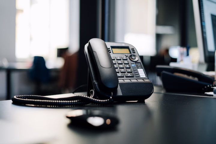 Telefono da ufficio su un tavolo, con cavo avvolto e computer sullo sfondo.